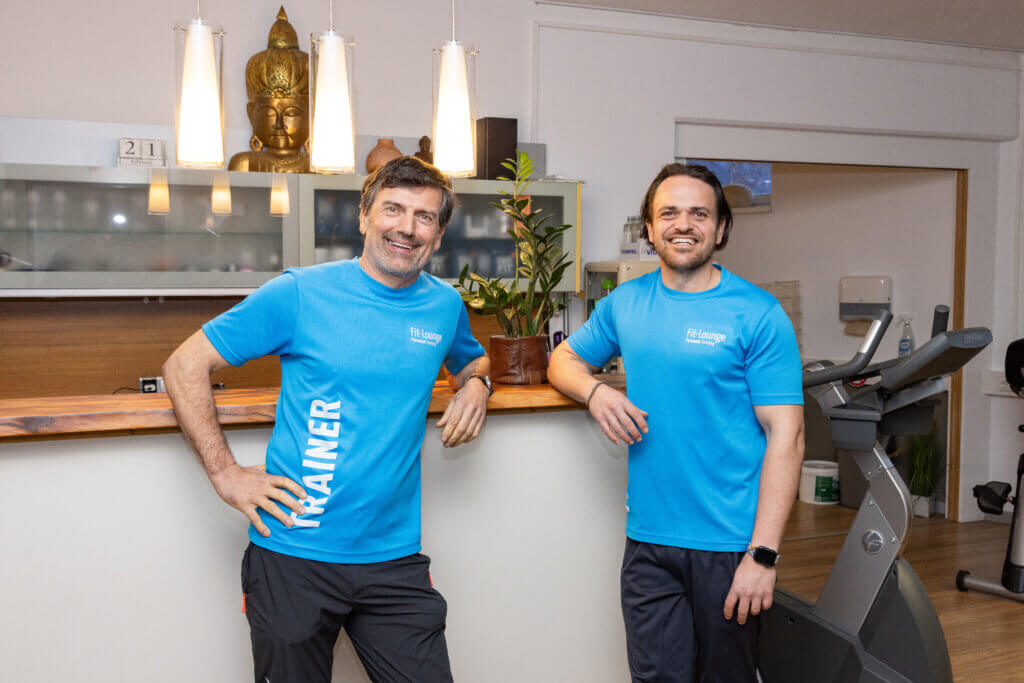 Zwei Trainer in blauen Shirts stehen an der Rezeption der Fit-Lounge in Bruck.